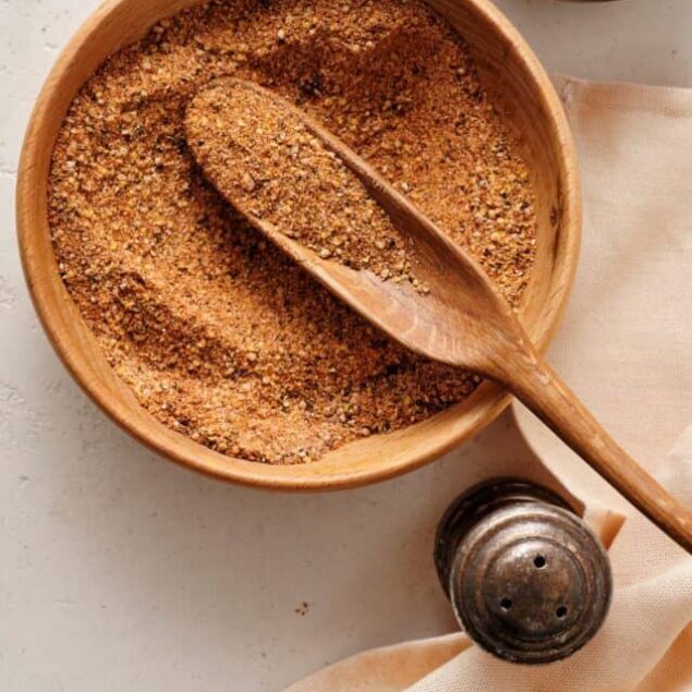 Ground spicy seasoning mixture in a wooden bowl with salt in a smaller bowl, on a neutral countertop.