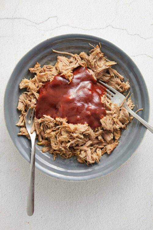 Shredded pork with barbecue sauce on a gray plate, two forks, on a textured white surface.