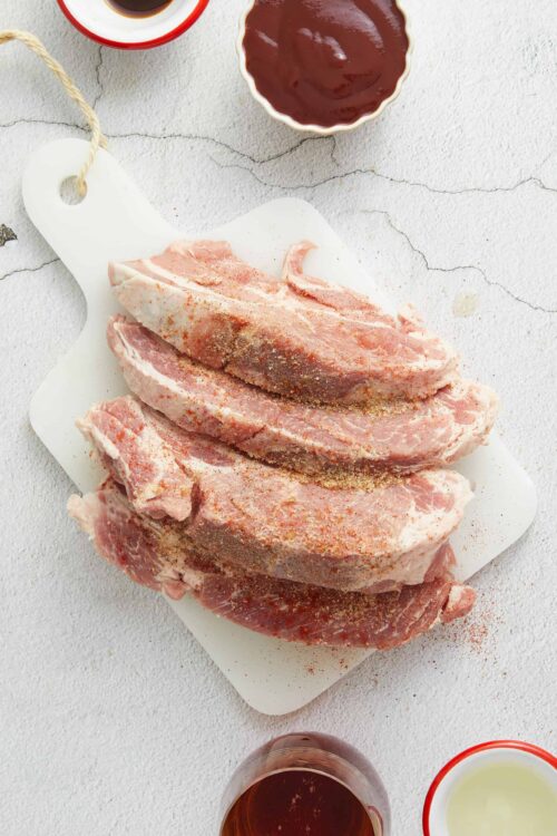 Juicy pork ribs seasoned with spices on white cutting board with barbecue sauces on white textured background.