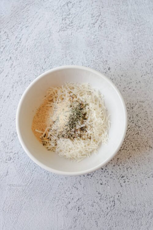 Shredded cheese in a white bowl with spices on top, ready for baking or cooking.