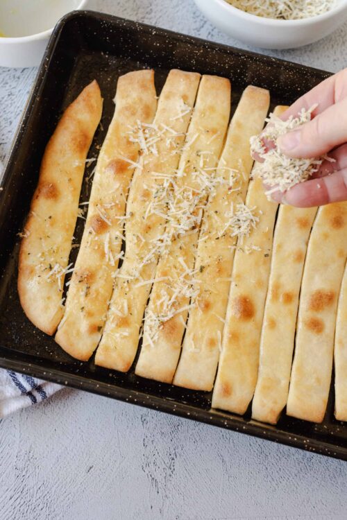Golden baked breadsticks topped with shredded cheese being added in a baking dish.