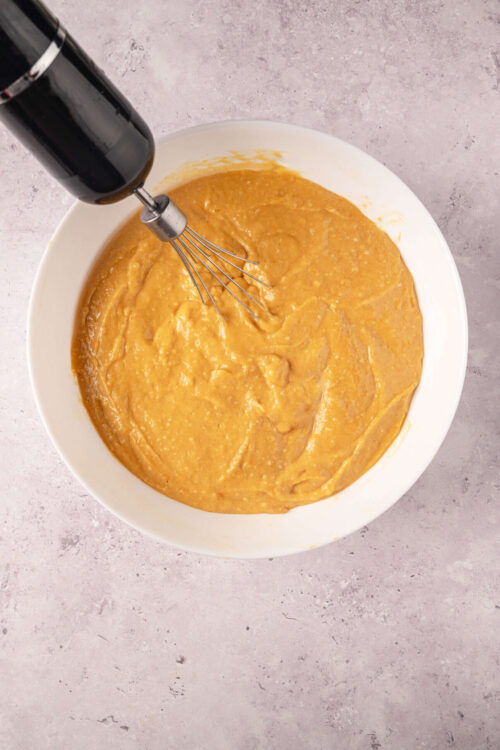 Creamy pumpkin pie filling with a hand mixer in a white bowl on a textured surface, ready for baking.