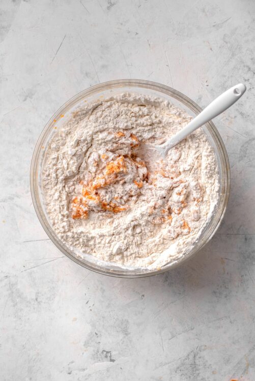 Flour mixture with orange zest and a white mixing spoon in a glass bowl for baking recipes.
