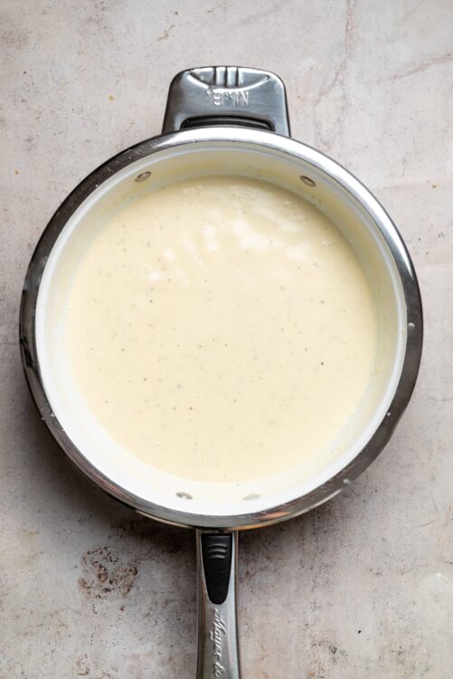 Creamy white béchamel sauce cooking in a stainless steel saucepan on a textured surface.
