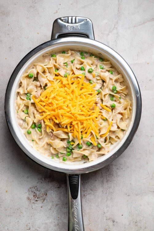 Creamy chicken and peas pasta casserole topped with shredded cheddar cheese in a stainless steel skillet.
