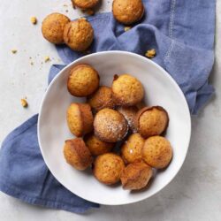 Golden vanilla cupcakes with lightly browned tops, arranged on a white plate with sugar and crumbs, on a light gray surface with a blue cloth.