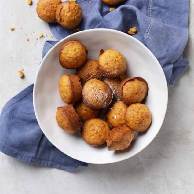 Golden vanilla cupcakes with lightly browned tops, arranged on a white plate with sugar and crumbs, on a light gray surface with a blue cloth.
