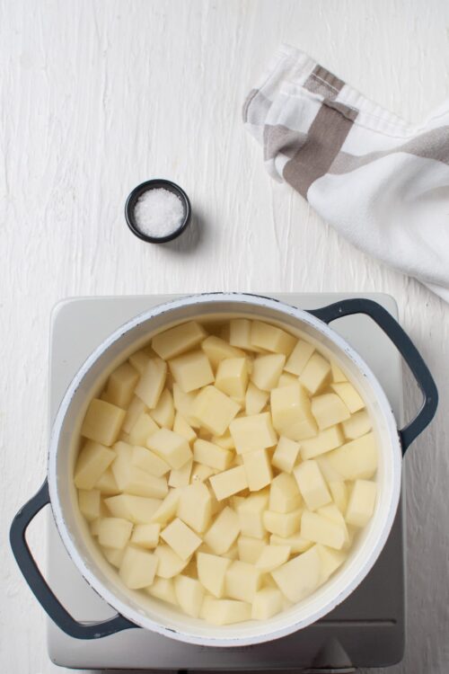 Cubed potatoes boiling in a pot on a stove with a salt shaker and kitchen towel nearby.