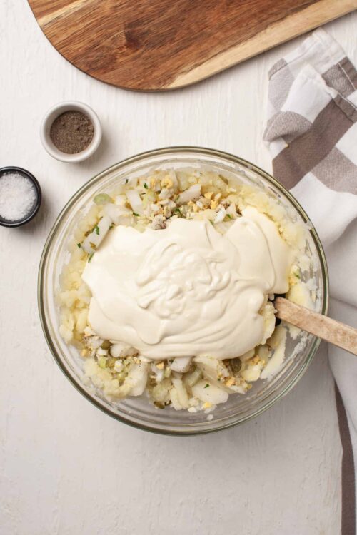 Creamy potato salad being prepared with mayonnaise, chopped potatoes, onions, and seasonings in a glass mixing bowl.