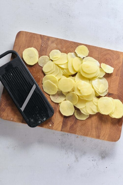 Thinly sliced potatoes on a wooden cutting board with a black mandoline slicer.