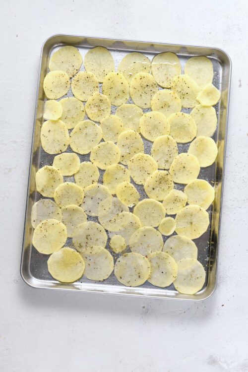 Thinly sliced potatoes seasoned with salt and pepper on a baking sheet.