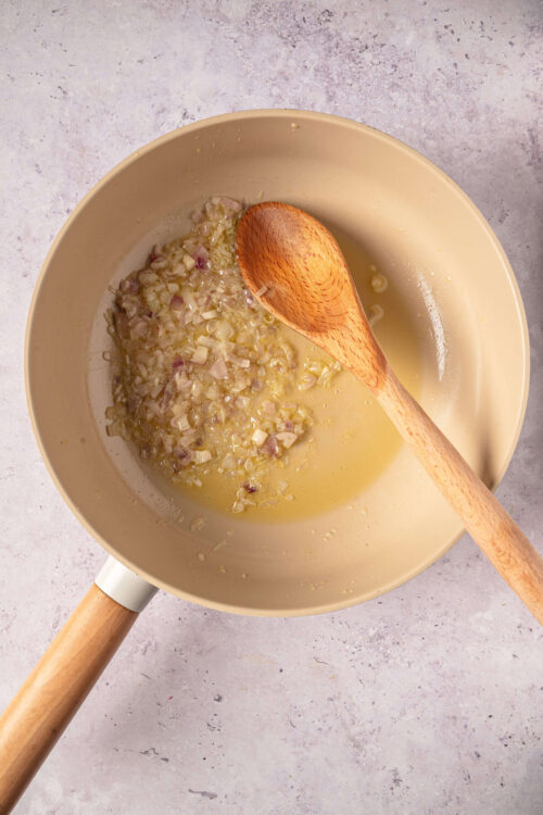 Sautéed onions cooking in a beige saucepan with a wooden spoon on a light textured surface.