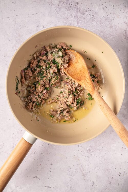 Sautéed ground beef in a beige skillet with chopped onions and herbs, with a wooden spoon, on a light countertop.