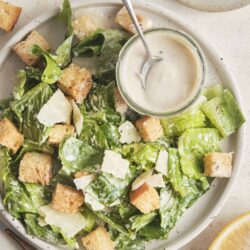 Fresh Caesar salad with croutons, parmesan, and creamy dressing on white plate, lemon wedge, salt and pepper, baked Bree recipe, easy healthy salad.