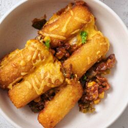 Crispy fried tacos with beef and cheese, served with savory topping, on white bowl for homemade Mexican-inspired dish.