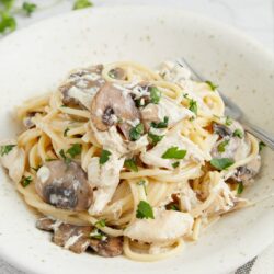 Creamy chicken mushroom pasta in white bowl, garnished with fresh herbs, on textured surface.