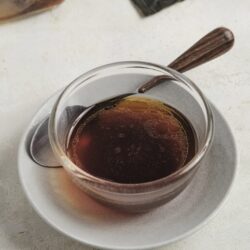 Rich brewed coffee in a glass cup with a metal spoon on a white plate, dark chocolate pieces and sugar jar in background.