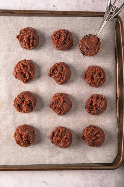 Gooey chocolate cookie dough balls on baking sheet with chocolate chunks and cookie scoop, no-bake chocolate cookies, bakedbree.com.