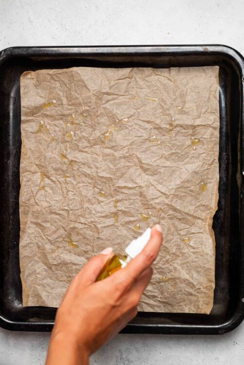 Olive oil being drizzled on parchment paper in a baking tray.