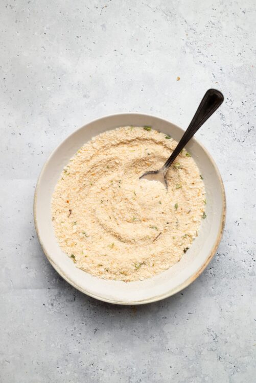 Breadcrumbs mixture with herbs in a white bowl on gray textured surface.