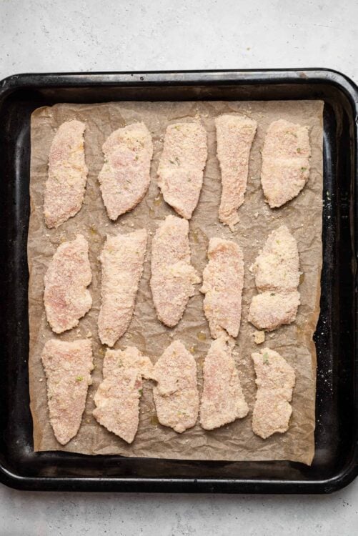 Breaded chicken tenders ready for baking, on a parchment-lined baking sheet.