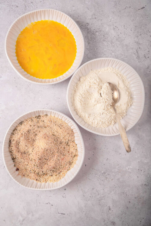 Flour, beaten eggs, and breading mix in white bowls for baking or frying.