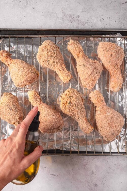 Crispy baked chicken drumsticks on a wire rack with aluminum foil, being sprayed with oil.