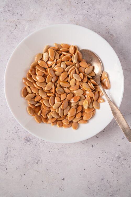 Roasted pumpkin seeds in a white bowl with a silver spoon on a light textured surface.