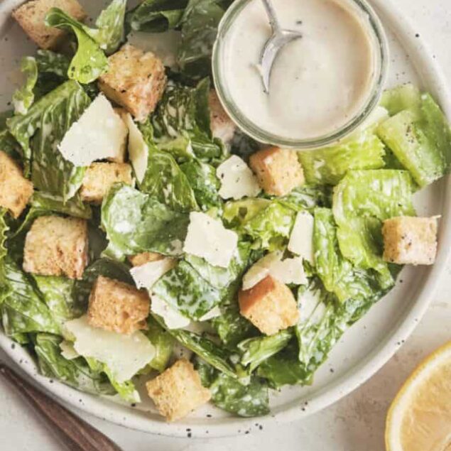Fresh Caesar salad with croutons, parmesan, and creamy dressing on white plate, lemon wedge, salt and pepper, baked Bree recipe, easy healthy salad.