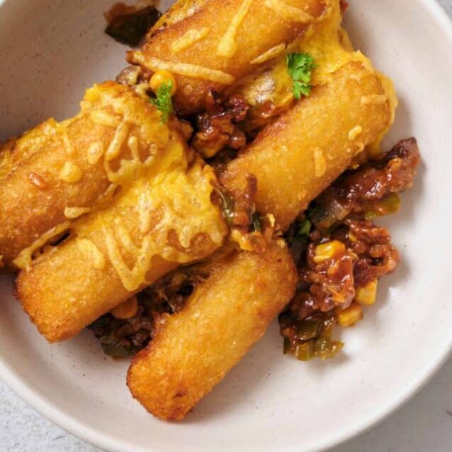 Crispy fried tacos with beef and cheese, served with savory topping, on white bowl for homemade Mexican-inspired dish.