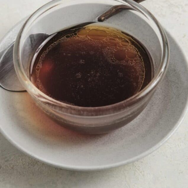 Rich brewed coffee in a glass cup with a metal spoon on a white plate, dark chocolate pieces and sugar jar in background.