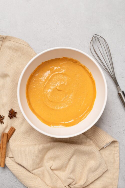 Creamy butternut squash soup in white bowl with a whisk on a light surface.