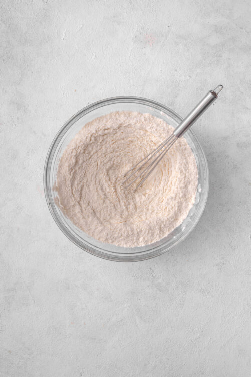 Flour mixture in glass bowl with whisk for baking recipes.