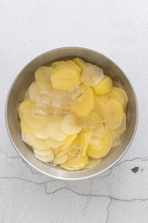 Thinly sliced potatoes with ice cubes soaking in water, ready for cooking or potato salad.
