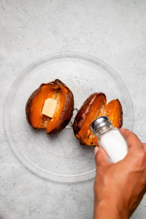Baked sweet potatoes being seasoned with salt on a glass plate.