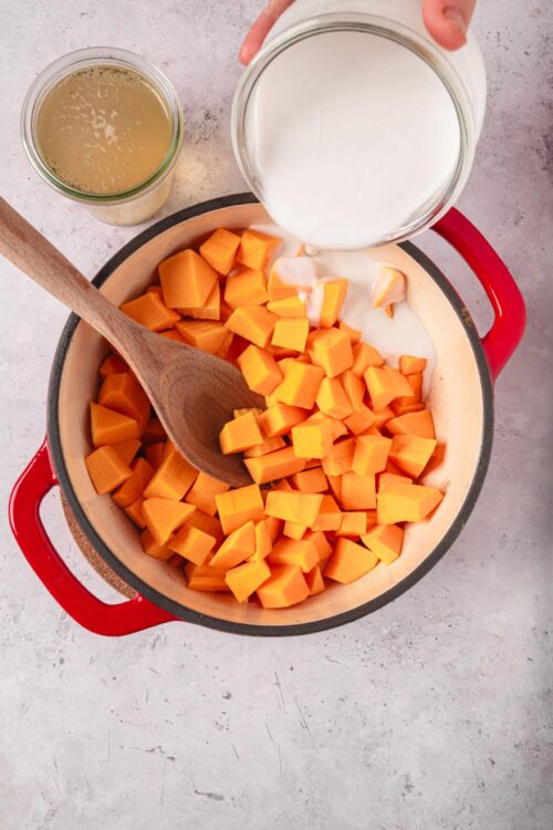 Creamy milk being poured over diced butternut squash in a Dutch oven for a baking or soup recipe.