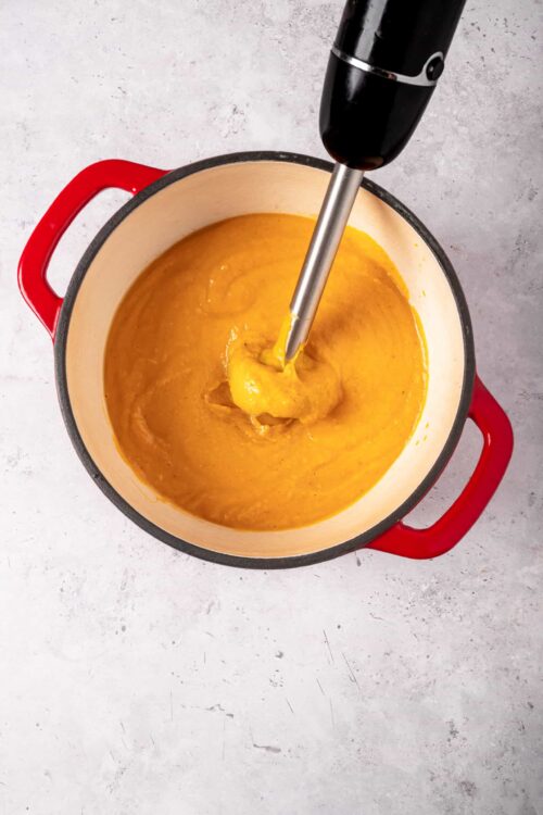 Creamy pumpkin soup being pureed with an immersion blender in a red Dutch oven.